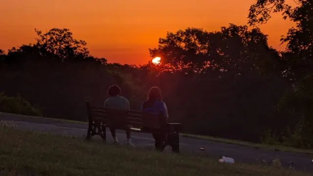 Two people sat at a bench in London, looking at the sunrise. The sky is bright orange in colour, with trees covering part of the sun.