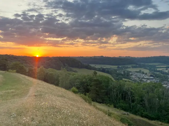 The sunrise coming over the trees to the left of the image. On the right, a village can be seen in the distance.