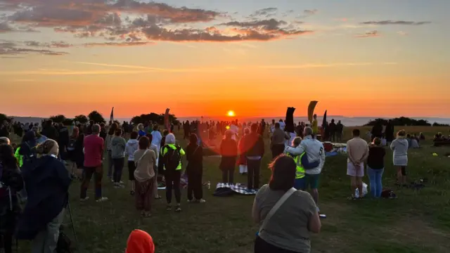 A few hundred people gathered in a field, looking at the sunrise in the distance.