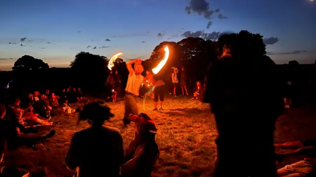 A circle of space surrounds a flame-thrower who is twirling a lit baton. A crowd of a few dozen people watch on from camping chairs and picnic blankets.