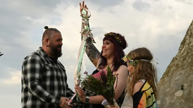 A man (L) in chequered shirt; a woman (C) in dress holding up some ribbons and flowers while she smiles; and a young girl (R) in front of the stones