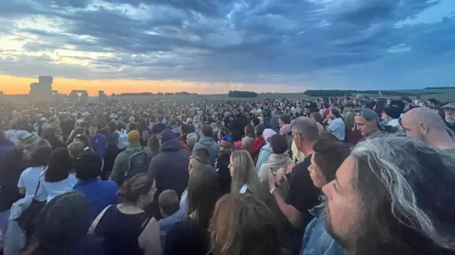 Thousands of people stand shoulder to shoulder as far as the eye can see. The stone circle at Stonehenge is visible in the distance. The sun is about to rise and has turned the sky partially orange.