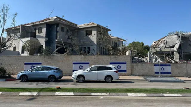 Debris and rubble is visible on a building that has been partly destroyed. Four Israeli flags are on a wall in front of the building. Two cars are parked in front of the wall.