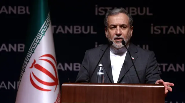 Iran Foreign Minister Abbas Araghchi delivers speech from behind a wooden lectern. He's wearing a dark grey suit with a white shirt, two mics in front of him, his left hand leaning on the lectern. An Iranian flag to his right