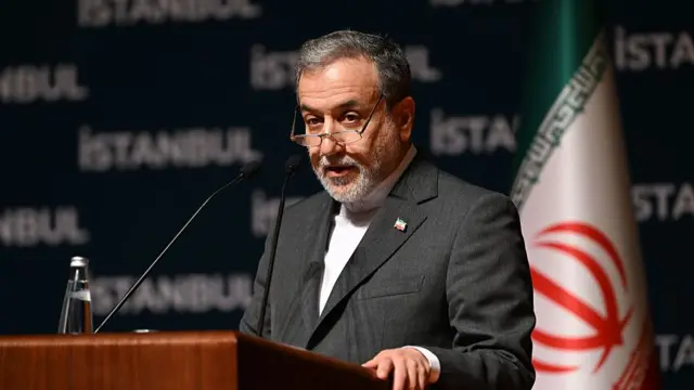 Iranian Foreign Minister Abbas Araghchi delivers speech behind wooden lectern. He's wearing a grey suit, white shirt as he stands on stage with an Iranian flag behind him