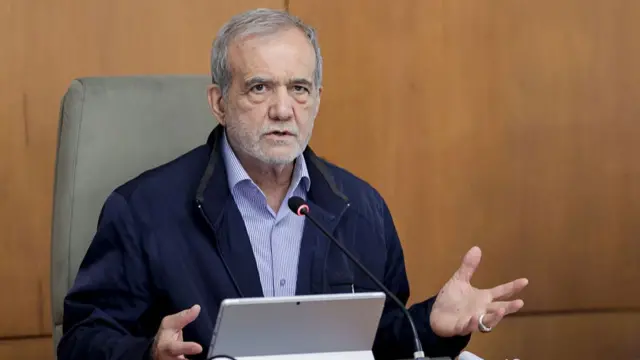 Iranian president Masoud Pezeshkian sitting down at a table with a tablet and table mic in front of him as he speaks. He's sitting in a high-back grey chair wearing a dark blue jacket and blue-striped shirt