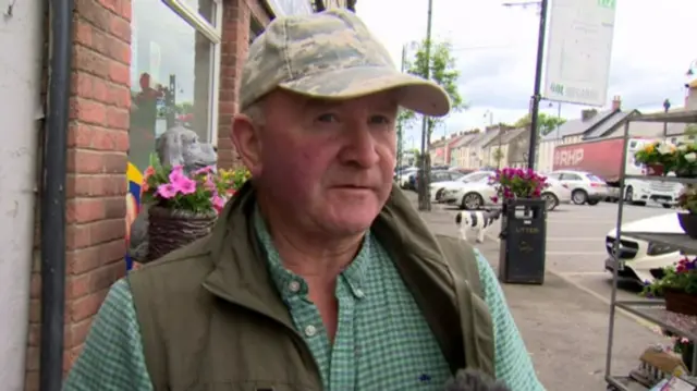 A man in a green chequered shirt wears a green Gillet and camo hat. He stands on the pavement of a high street.