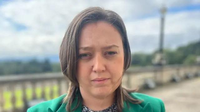 Deborah Erskine pictured outside Stormont last year.  She has shoulder-length brown hair and is wearing a bright green suit jacket over a ditsy floral print top or dress