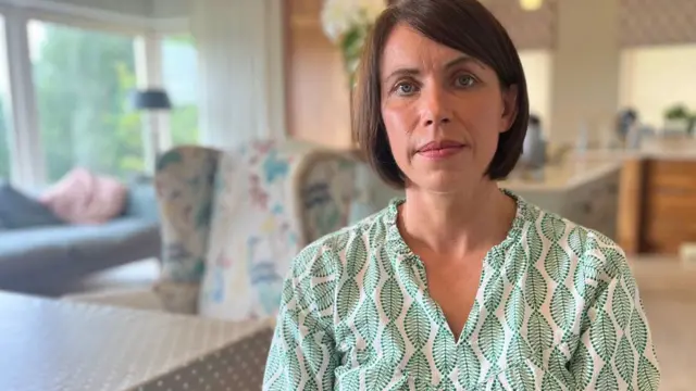 A woman is looking into the camera. She's sitting in a living room. She's wearing a green and cream top and has shirt brown hair.