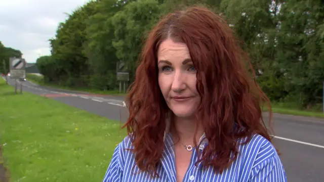 Emma is looking away from the camera. She has curly hair and is wearing a stripped blouse. She's standing by a road.