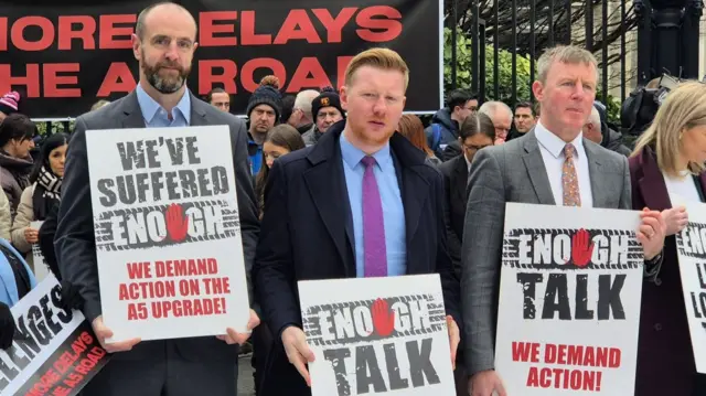 A group of campaigners are standing together holding banners.  Some state, enough is enough, while others state we've suffered enough.