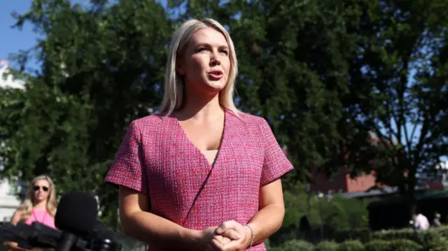 Leavitt, in a pink dress, clasps her hands as she speaks outside under a tree