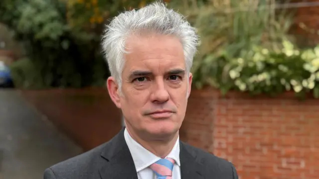 James Cartlidge stares directly at the camera and wears a dark grey blazer white shirt and salmon and baby blue striped tie