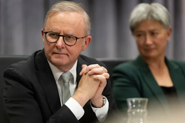 Australia's Prime Minister Anthony Albanese, seated, in a suit and glasses, with hands clasped and looking to left, with Foreign Minister Penny Wong, blurred, in background on right