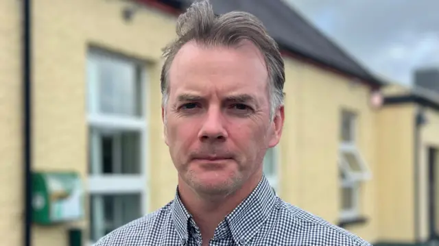 A man with thinning brown-grey hair and a black and white check shirt looks into the camera. He is standing outside a yellow building. It's an overcast day.
