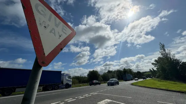 A5 road with Give Way sign in foreground. There is a lorry and cars on the road. The sky is blue.