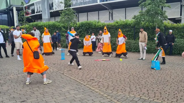 A group of people, most of whom are in traffic cone fancy dress, play cricket outside the stands at Headingley
