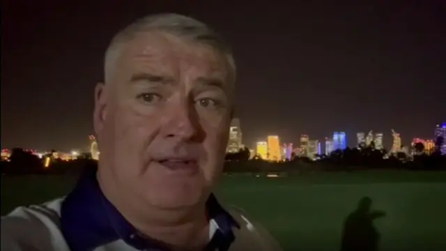Rhodri Ogwen Williams speaks to the camera, where behind him the Doha skyline is lit up at night