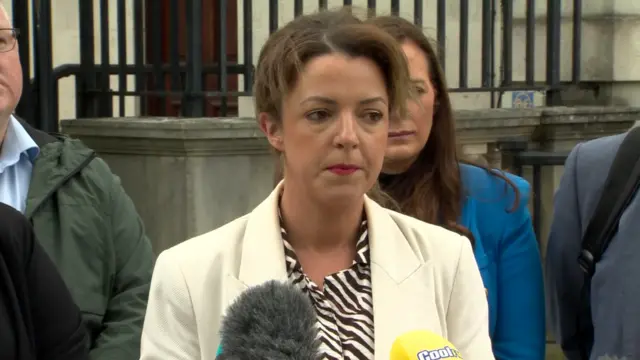 Liz Kimmins speaking to reporters outside court.  She has shoulder-length brown hair tied back and she is wearing a white jacket over a black and white blouse.
