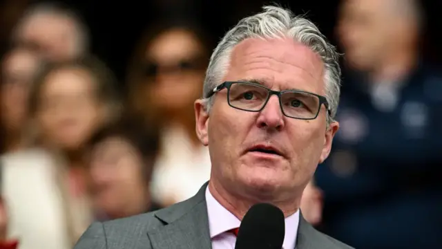 A man holding a microphone up to his mouth, he is wearing glasses, a grey suit and pink shirt and has short grey hair