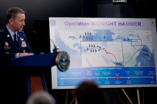 A Pentagon official, blurred to the far left and in uniform, speaking at a podium with a large poster titled Operation Midnight Hammer with a graphic detailing US strikes in Iran