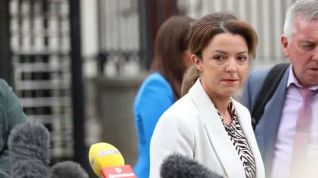 A woman in a white suit and balck and white striped top stands away from four microphones. She has long brown hair. Behind her is a grey haired man in a grey suit, pink shirt and pink tie.