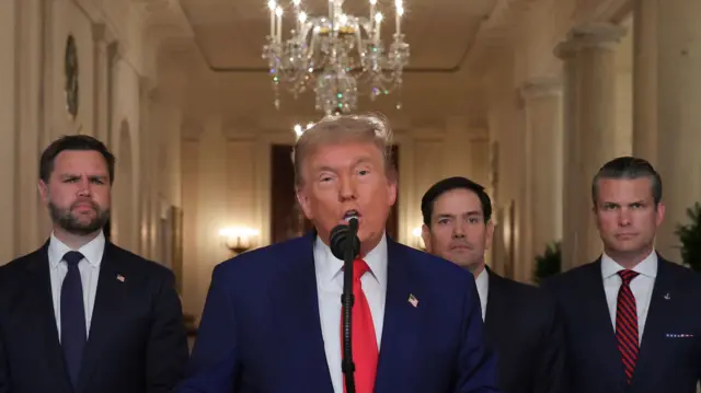 Trump speaks into a microphone below a chandelier with US officials around him.