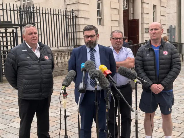 Niall McKenna, chair of the Enough is Enough campaign group, speaking to the the media outside court.  He is wearing a navy suit and has short, dark hair, a dark beard and glasses. Three other men are flanking him as he stands at the microphones