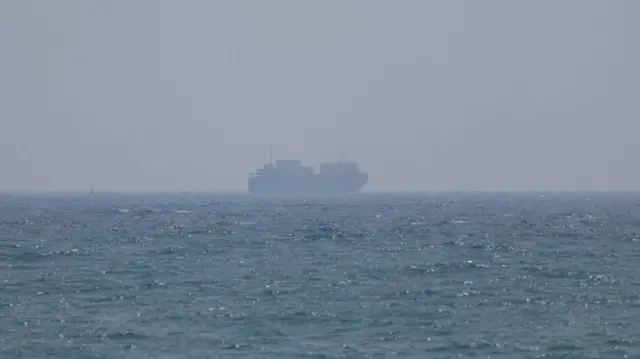 A container ship seen in the distance across the sea. The sky is blue but looks hazy