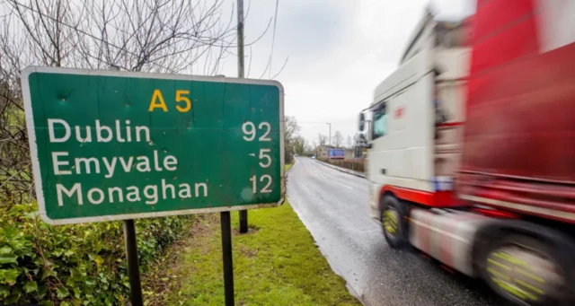 A photo of a red and white lorry driving past a green road sign on the A5.  The sign says "A5 - Dublin 92 (miles) Emyvale 5 (miles) Monaghan 12 (mile)