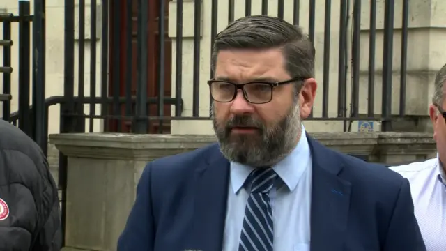 Niall McKenna standing outside the court. He has short dair hair and a dark but greying beard.  He is wearing glasses and a navy suit.