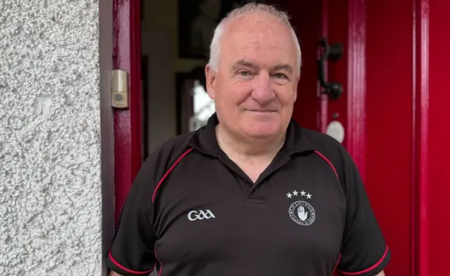 Jim Hunter has grey balding hair, he is wearing a black polo with red piping - there is a tyrone crest on his left chest and GAA on the right