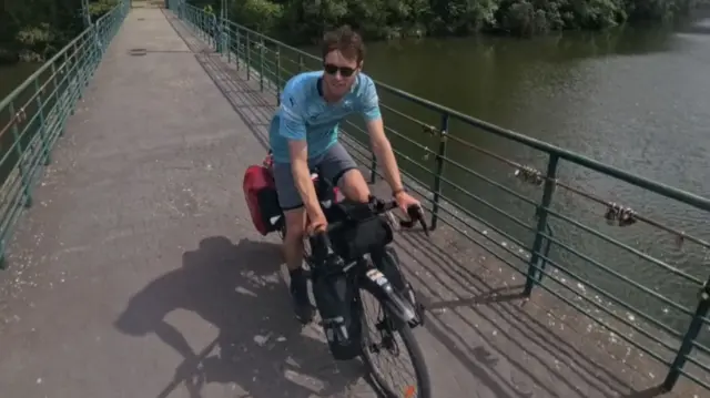 Ollie, wearing a blue shirt and dark sunglasses, cycles on a bridge over a river.