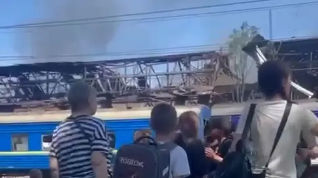 A blue train is seen in front of a damaged building with some rising in the distance