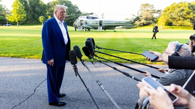 US President Donald Trump speaks to the media as he departs the White House for the NATO leaders summit, TV cameras and microphones can be seen with the Marine One helicopter in the distance on the White House lawn