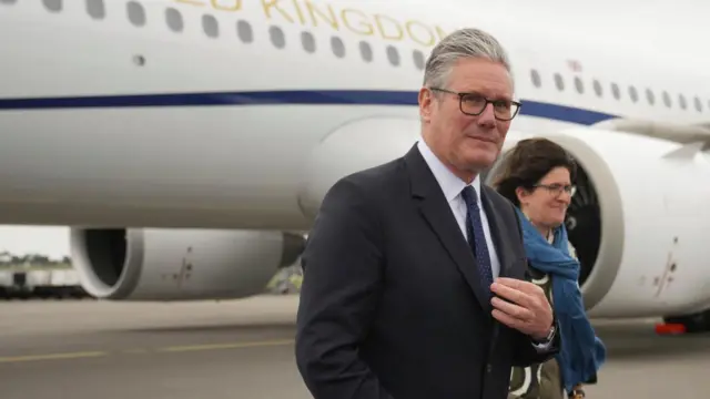Keir Starmer looks to the camera and touches his tie as he walks on the tarmac on arrival in the Netherlands. There is a plane in the background.