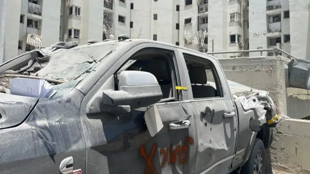 A big pick up truck sits idle with its windows blown out and dust scattered over it - following a missile strike in Beersheba. A tower block behind it also has many of its windows blown out