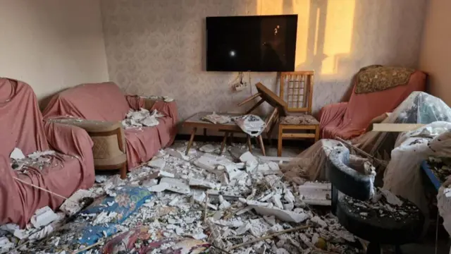 Debris scattered over and across a living room with sofa's covered in broken bits of a building