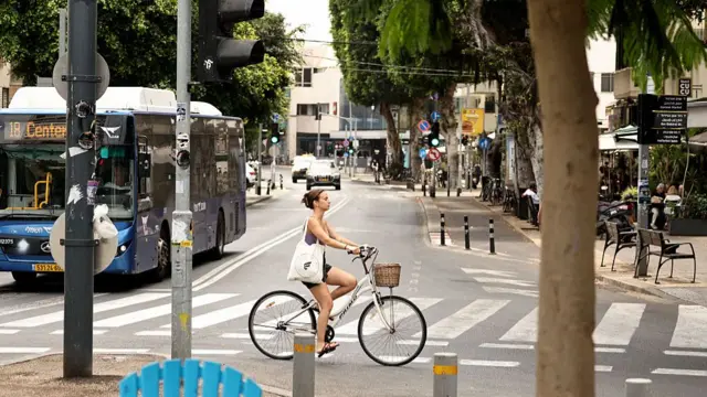 A woman rides her bike on a street - cafes are open in the background, and a bus is driving down the street