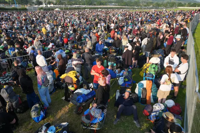 Queues of people are snaking round metal barriers to get into Glastonbury Festival. It is a sunny day and people are wearing hats, shorts and bum bags.