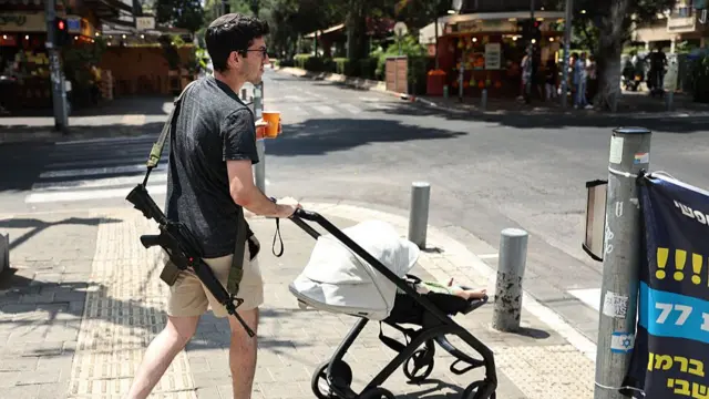 An armed Israeli pushing a pram walks down a street in Tel Aviv