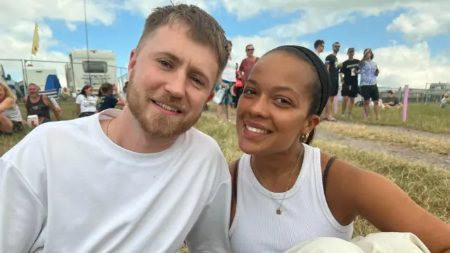 Will Laing and Aaliyah Howard sat on the ground. Behind them you can see two caravans and a group of men standing up.