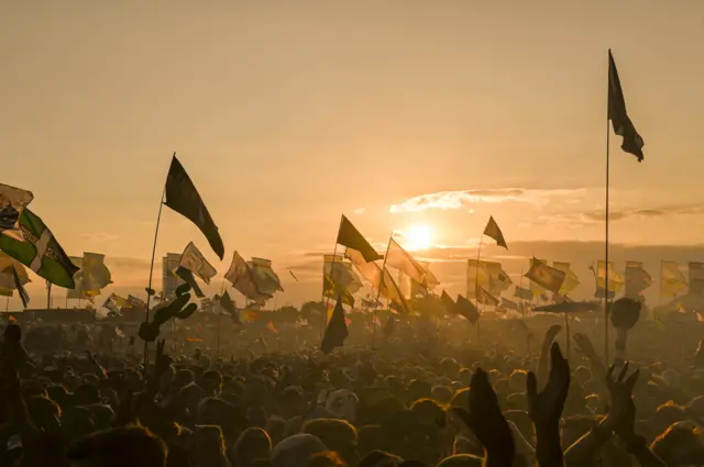 A hazy orange-tinged picture of a huge crowd at Glastonbury, showing people with their hands in the air and waving flags.