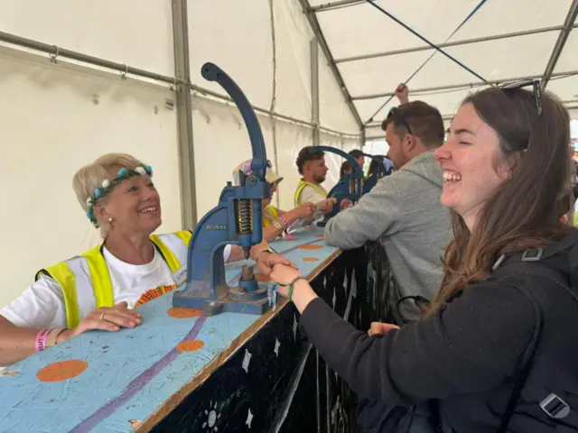 A woman in a floral headband and high-vis vest smiles as she clamps a wristband onto the wrist of a laughing woman.