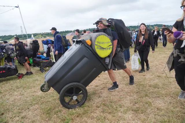 A man in a bucket hat pushes a huge black wheely bin full of camping equipment across the grass.