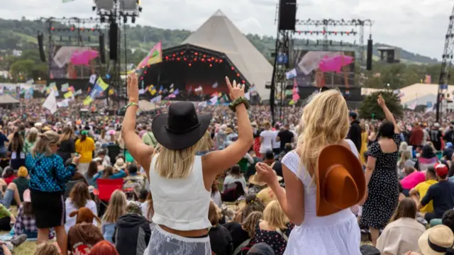 Two girls in the foreground (L: wearing a dark brown cowboy hat, white tank top and black skirt covered by white lace sarong. She has her hands raised. The other girl, R, is in a white sleeveless dress with a light brown cowboy hat hanging down her back). In front of them, a large crowd looking performers on the Pyramid Stage at Glastonbury