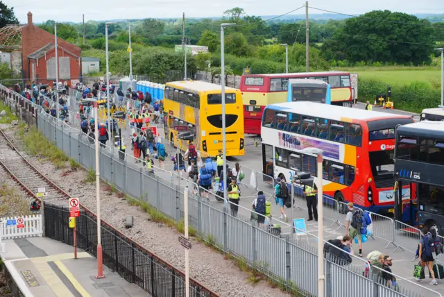 Five shuttle buses parked next to Castle Cary station, with festivalgoers seen carrying large backpacks past metal bollards.