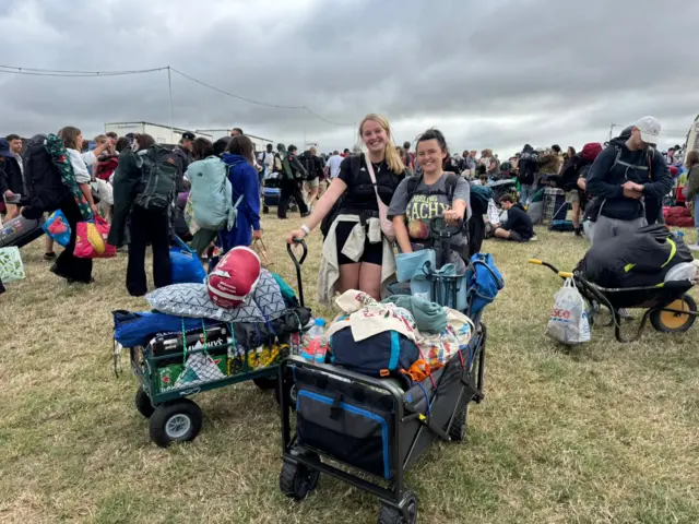 Annabel and Louise smile and pose with large trolleys packed with their camping equipment.