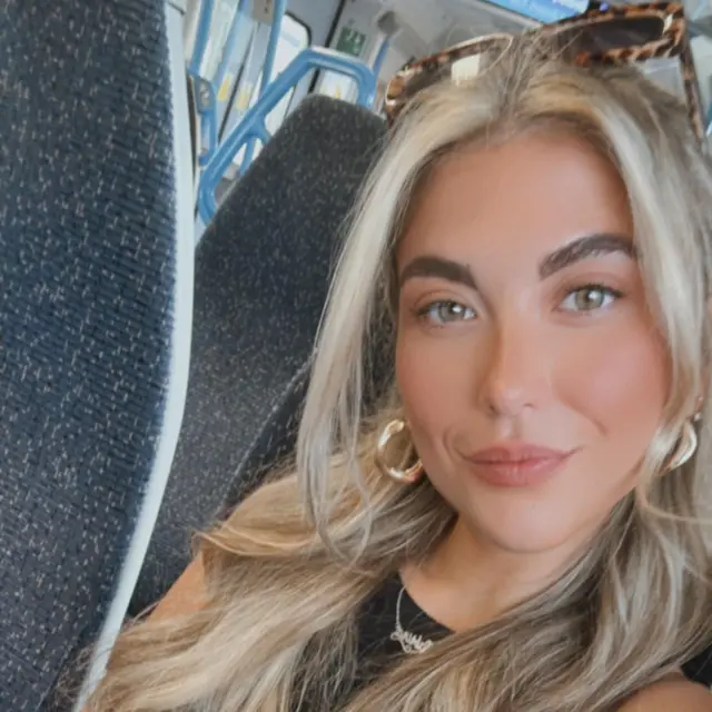 A woman with blonde hair looks into camera, while sat on a bus