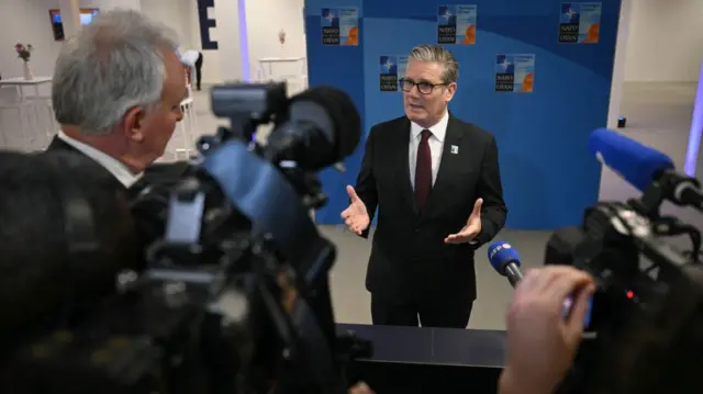 Keir Starmer standing in front of a grey-haired journalist (L, with his back to the camera) as he answers questions upon arriving to the Nato summit in The Hague. They're indoors and Starmer is standing in front of a blue wall with four Nato logos